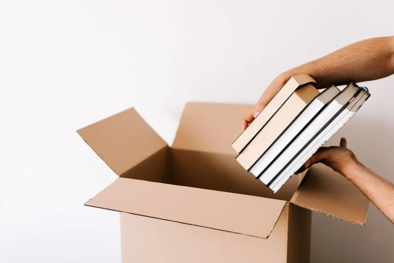 Deux mains posent une pile de livres au fond d’un carton ouvert